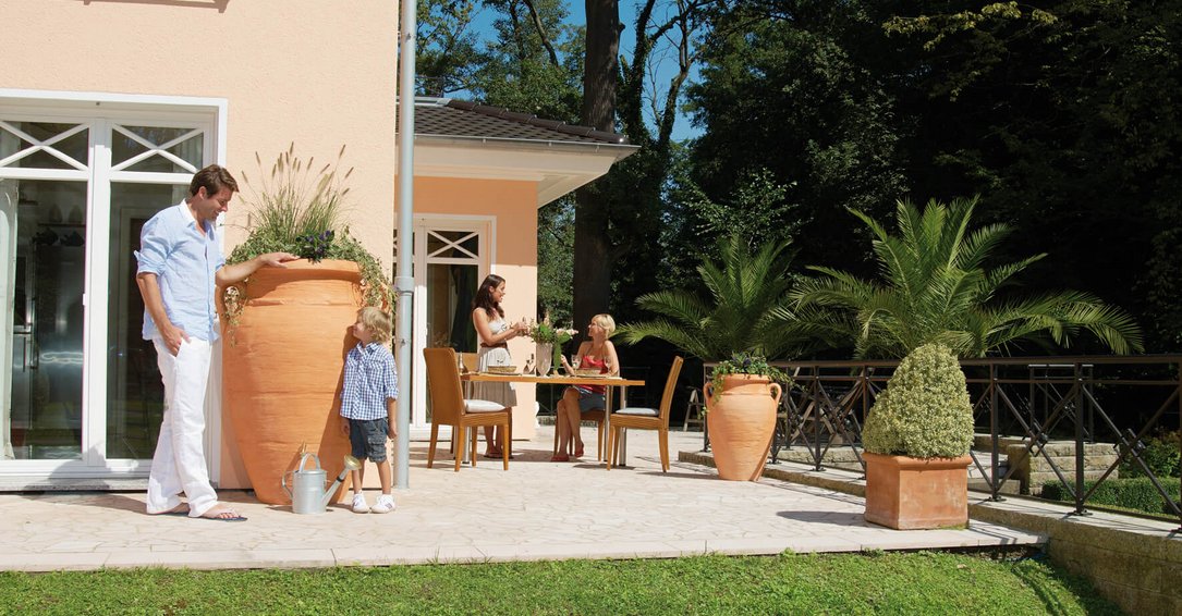 une famille sur sa terrasse au soleil avec le père et l'enfant au pied d'une cuve aérienne de récupération d'eau de pluie Amphore Antik Terracotta de Sotra'O dans l'Oise