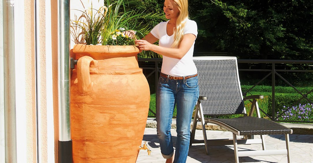 une femme s'occupe des fleurs du bac intégré de la cuve aérienne amphore antik terracotta Sotra'O de récupération des eaux de pluie dans son jardin dans les hauts-de-france